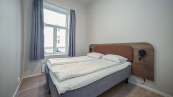Un llit individual amb capçalera de fusta, una finestra amb cortines i una làmpada penjant a la paret.