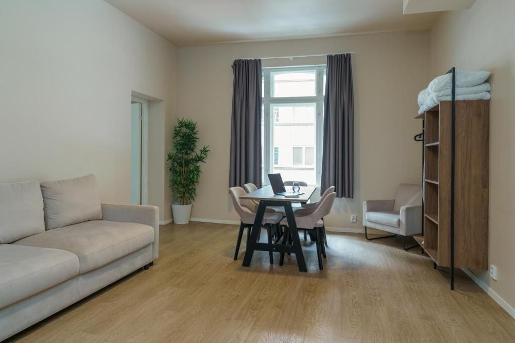 Una sala de estar con un sofá, una mesa de comedor con sillas, una planta en maceta, una ventana con cortinas y una estantería de madera.