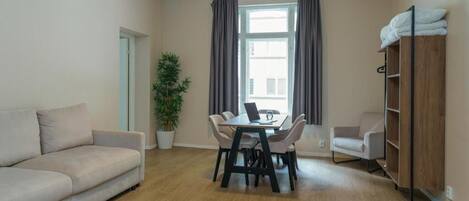 Una sala de estar con un sofá, una mesa de comedor con sillas, una planta en maceta, una ventana con cortinas y una estantería de madera.