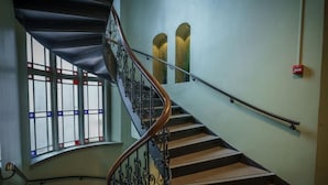 Una escalera de caracol con barandilla de madera y balaustradas de metal negro, que conduce a un rellano con un ventanal curvo con vidrieras.