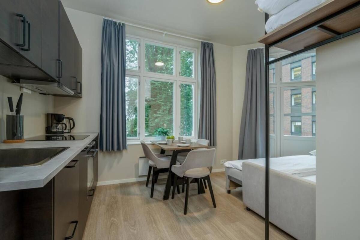 Ein kompaktes Hotelzimmer mit Küchenzeile, Essbereich, Bett und großem Fenster mit Vorhängen.
