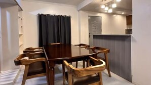 A dining area with a wooden table and chairs, a kitchen area with a refrigerator and cabinets, and a ceiling light fixture. - Balay Tori & Gil (Baguio)