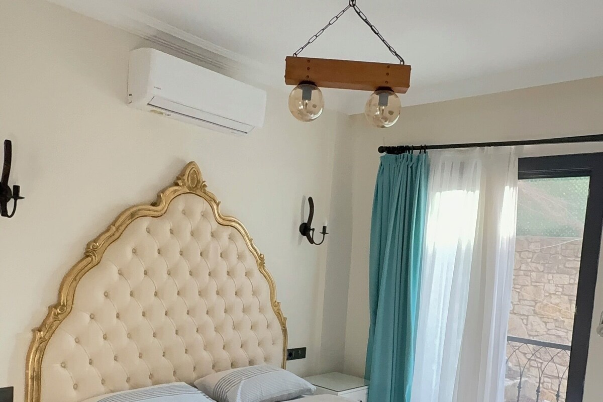 A bedroom with a tufted headboard, a bed with white linens, a bedside table, a hanging light fixture, and a view through a window with curtains.