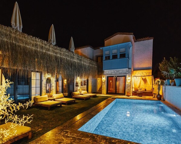 A poolside area with lounge chairs, a thatched roof, and a building with a blue facade. - Alaçatı Paradise Hotel (Cesme)