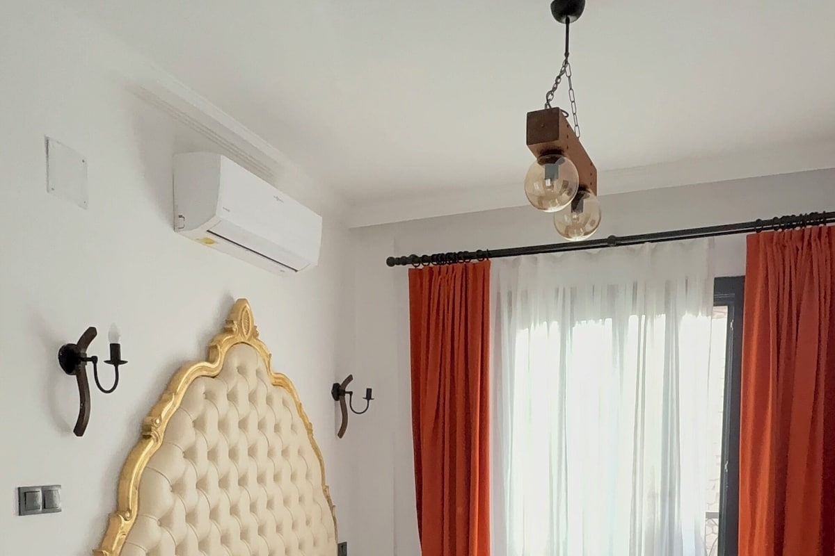 A bedroom with a bed, bedside tables, an air conditioning unit, a ceiling fan, and curtains.