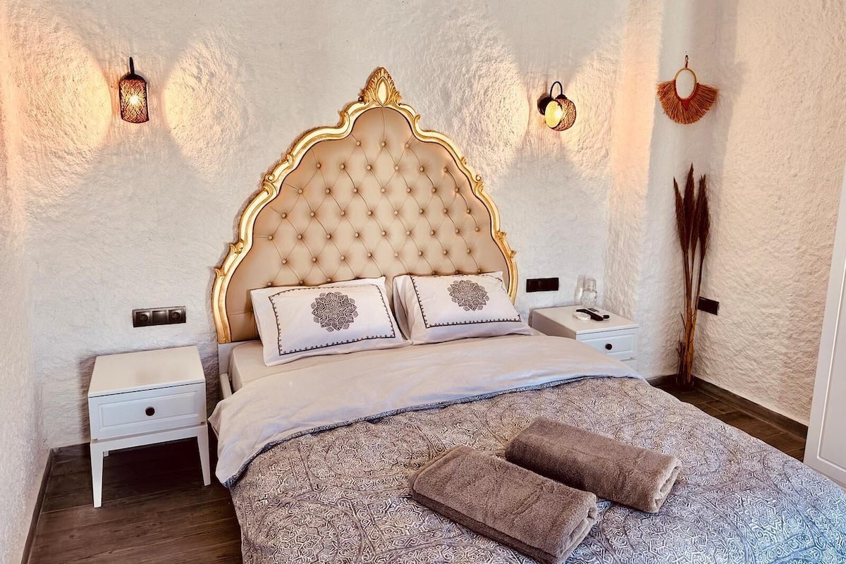 A bedroom with a tufted headboard, two bedside tables, a bed with patterned bedding, and decorative wall sconces.
