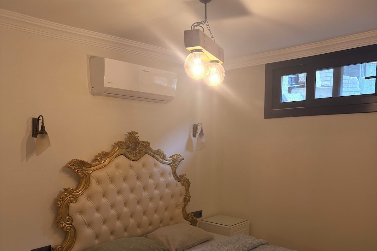 A bedroom with a tufted headboard, a bedside table with a lamp, a nightstand, a window with a view, and an air conditioning unit.