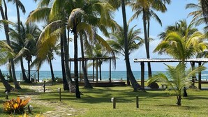A tropical garden with palm trees, a wooden deck, and a view of the ocean. - Beachfront Apartment on the Upper Floor: Best Value for Money (Maraú)
