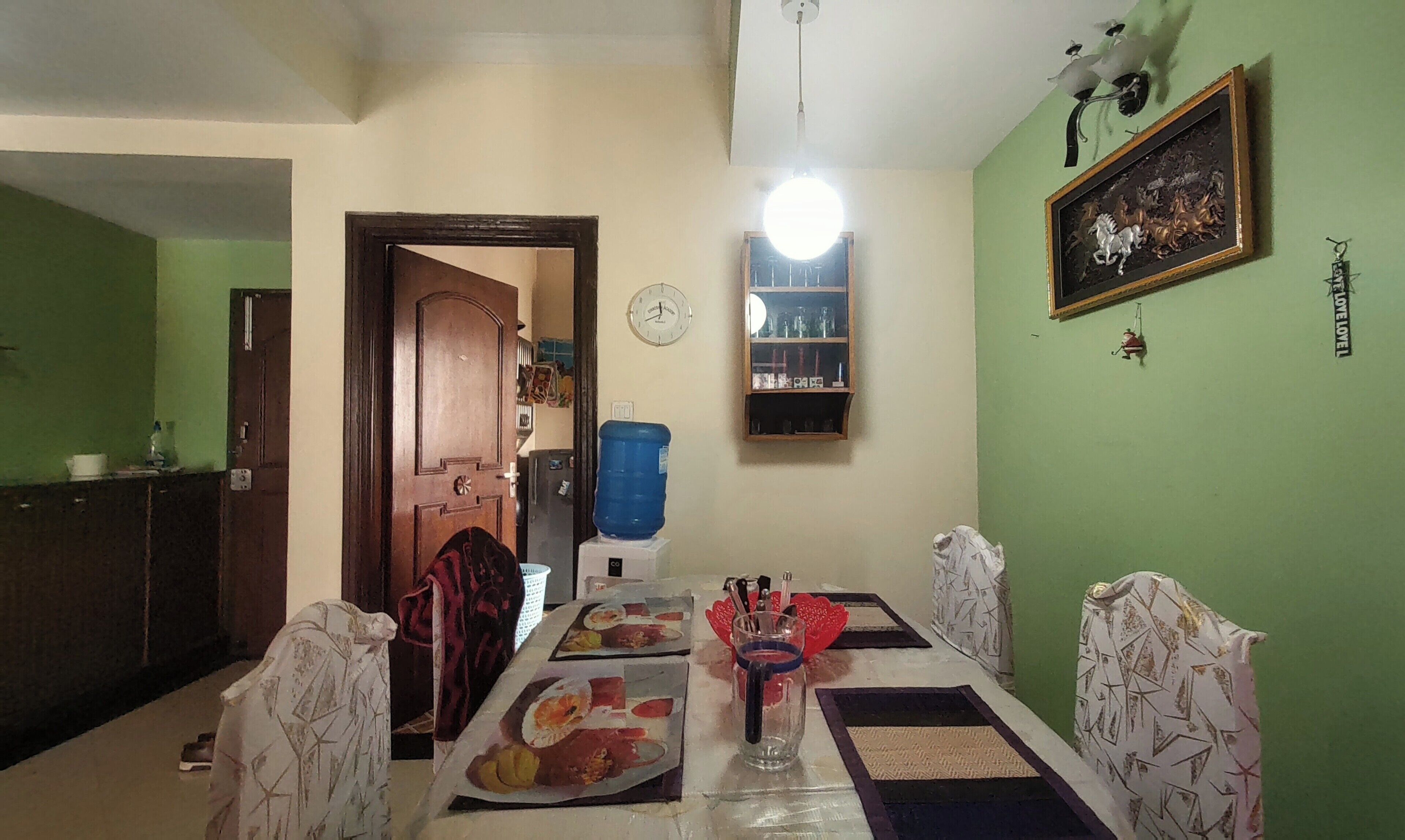 A dining area with a table set for two, chairs, a water dispenser, a clock, and a framed picture on the wall.