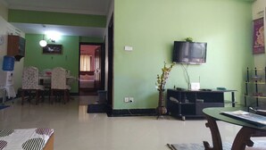 A living room with a green wall, a television mounted above a black stand, a dining area with a table and chairs, and a hallway leading to another room. - Metro axis residency (Kathmandu)