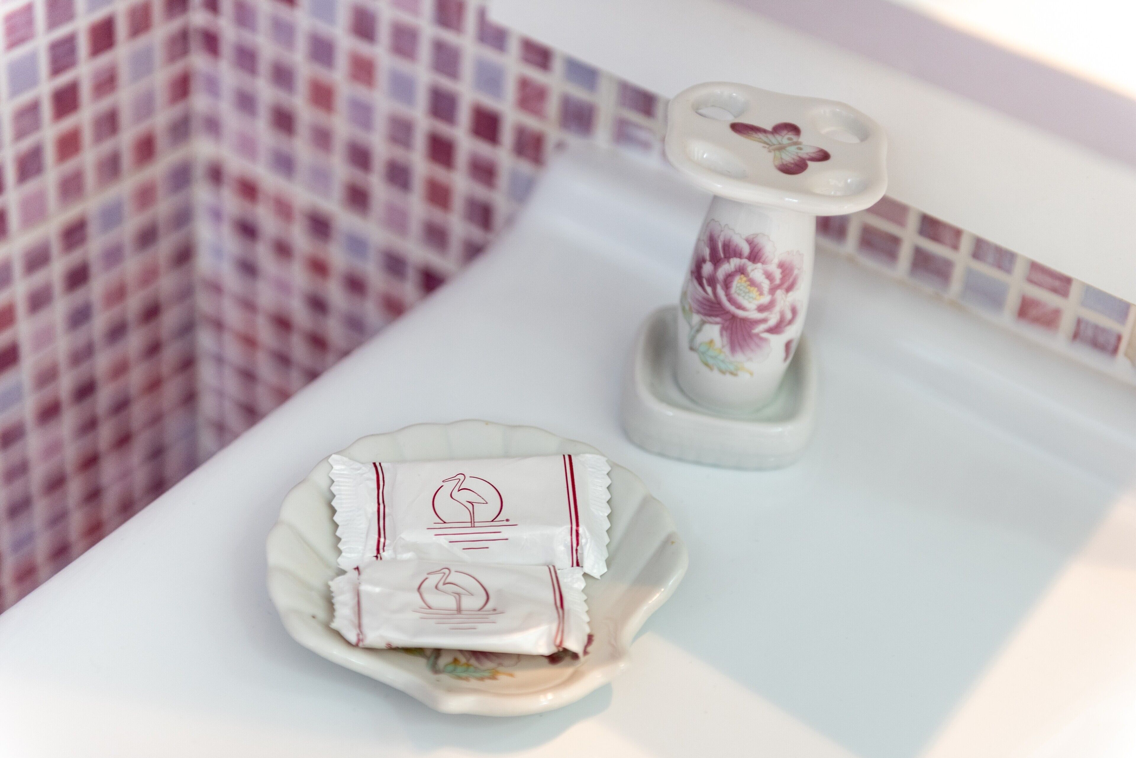 Une salle de bain avec un mur carrelé, un lavabo blanc, un vase décoratif et une assiette contenant deux objets emballés.