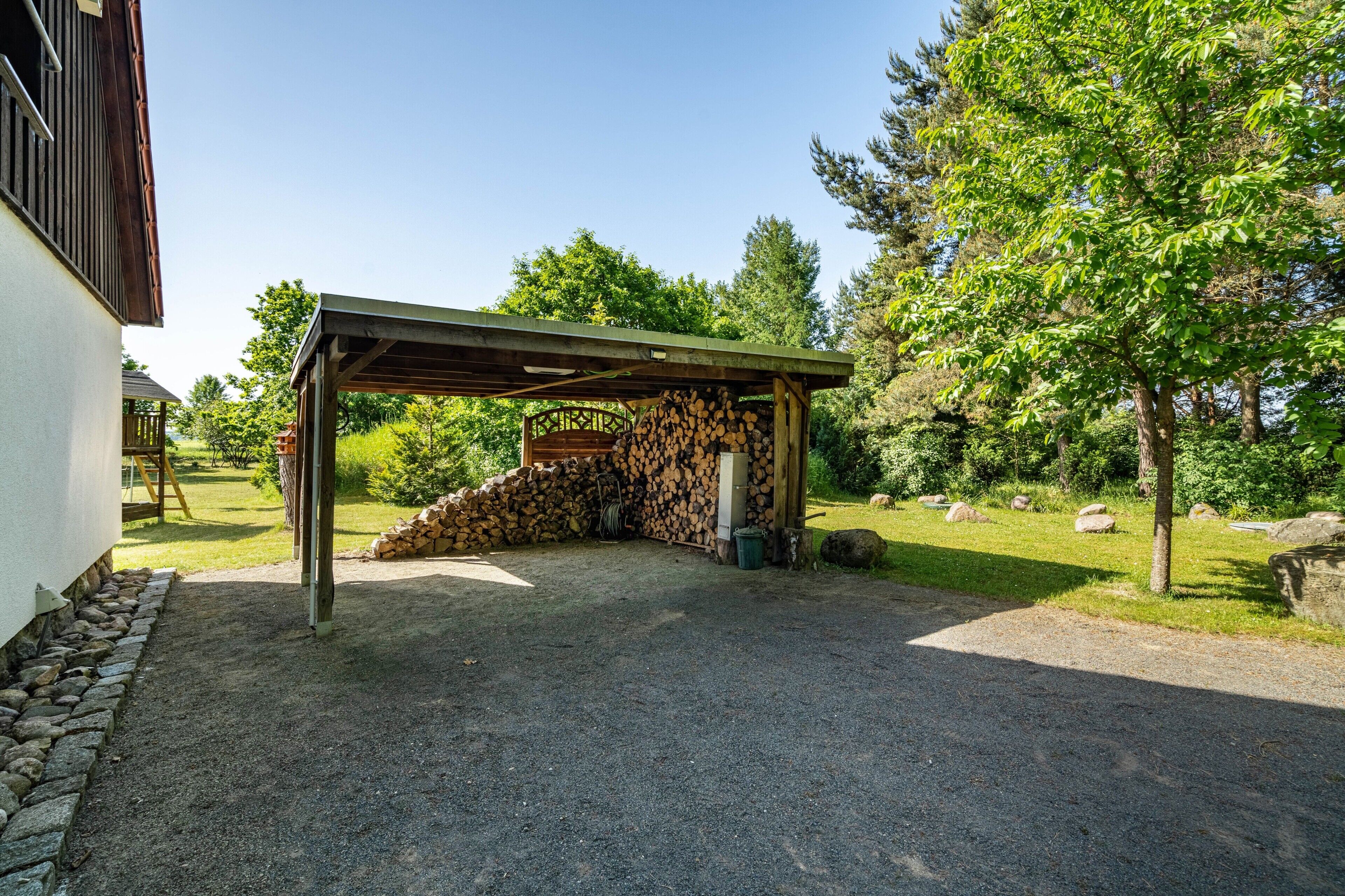 A covered outdoor area with a stack of firewood, a stone wall, and a gravel driveway.