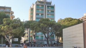 A sandy beach with a tall residential building in the background, bicycles parked near a building, and a clear sky. - Sea-front Accommodation in Lignano Sabbiadoro (Lignano Sabbiadoro)