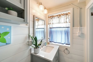 Casa de banho com lavatório branco, espelho, planta em vaso e janela com cortinas.