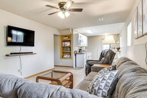 Un salon avec un canapé, une table basse, un téléviseur, un ventilateur de plafond et, au fond, un coin cuisine.