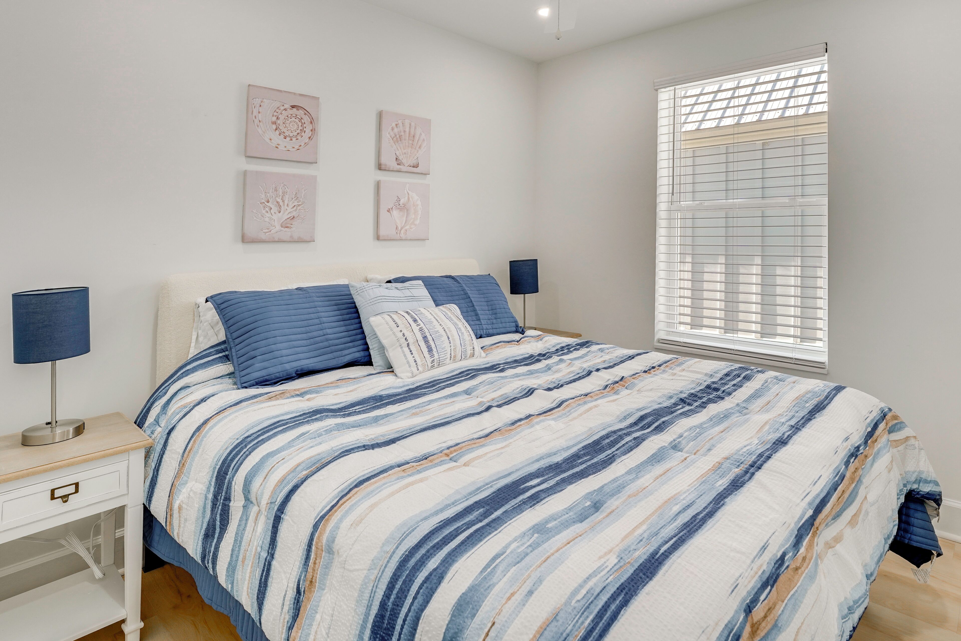 A bedroom with a bed, bedside tables, wall art, and a window with blinds.