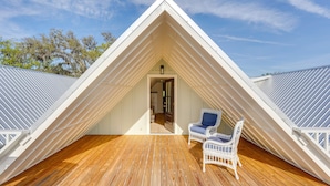 Una habitación ático triangular con una terraza de madera, dos sillas de mimbre y una puerta que da a un espacio interior.