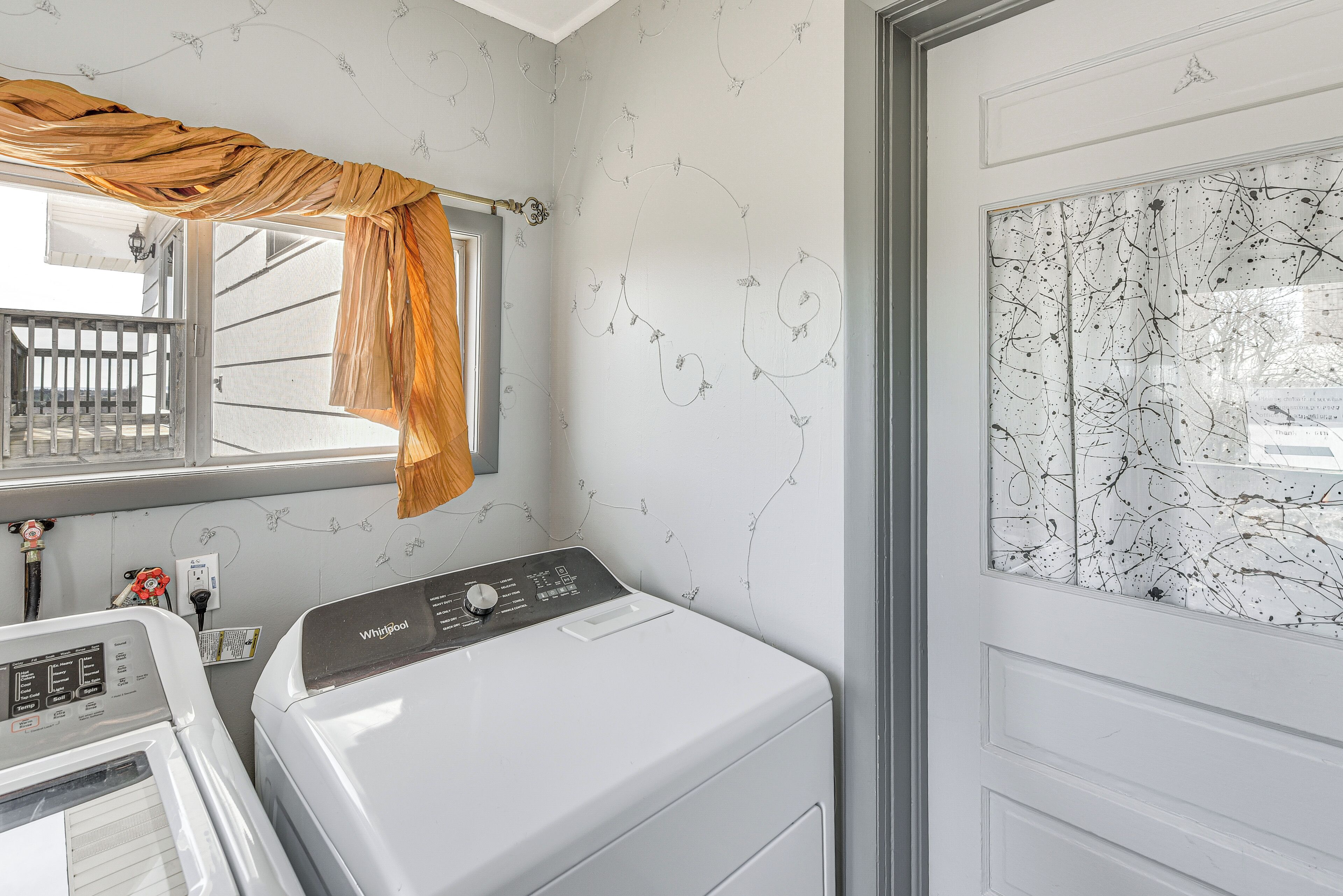 A laundry room with a front-loading washer and dryer, a window with a curtain, and a wall with a floral pattern.