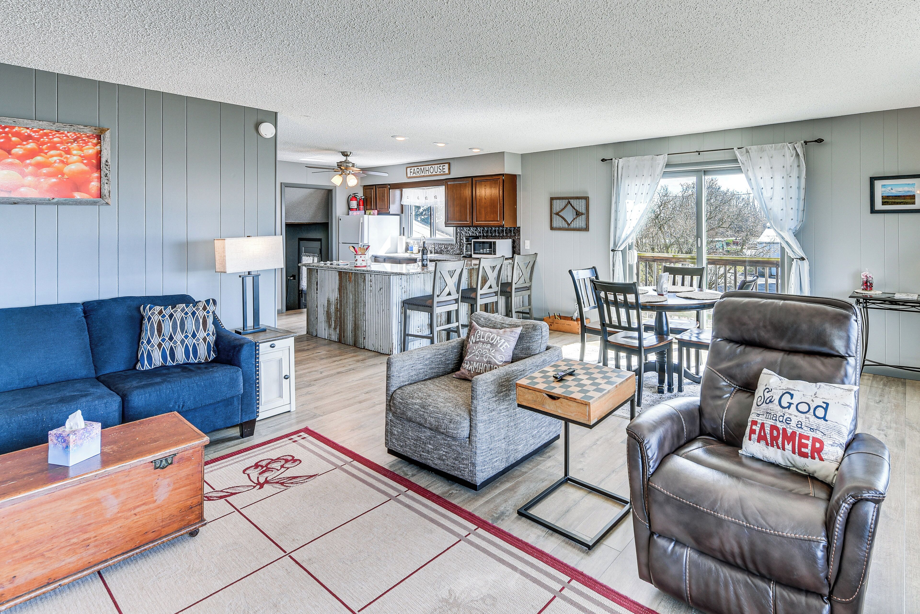 A living room with a sofa, armchair, and coffee table, a kitchen in the background, and a view through the window.