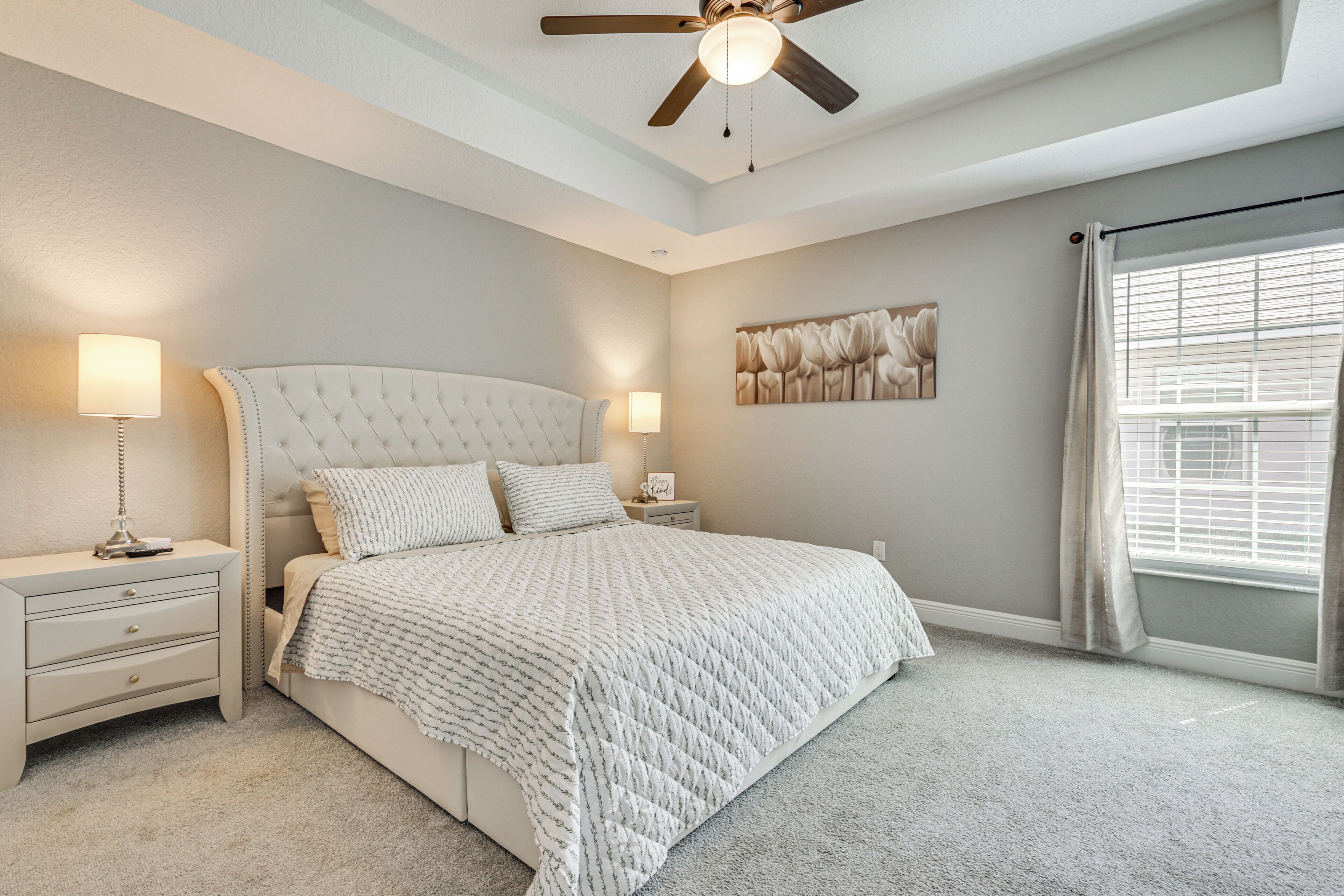 A bedroom with a tufted headboard, a large bed with a quilted bedspread, two bedside tables with lamps, a ceiling fan, and a window with blinds.