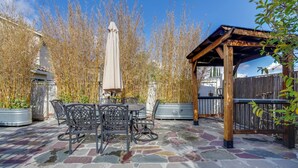 Una terrazza esterna con pavimento in pietra, tavoli e sedie in metallo, una pergola e piante alte.