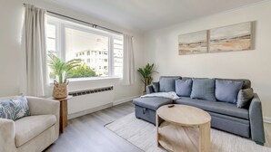A living room with a grey sofa, a beige armchair, a round wooden coffee table, a large window with curtains, and two wall-mounted paintings.
