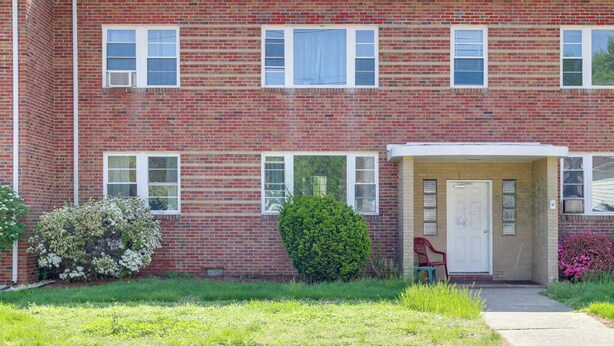 A two-story brick building with a front porch, a door, and a small garden area with flowers and shrubs.