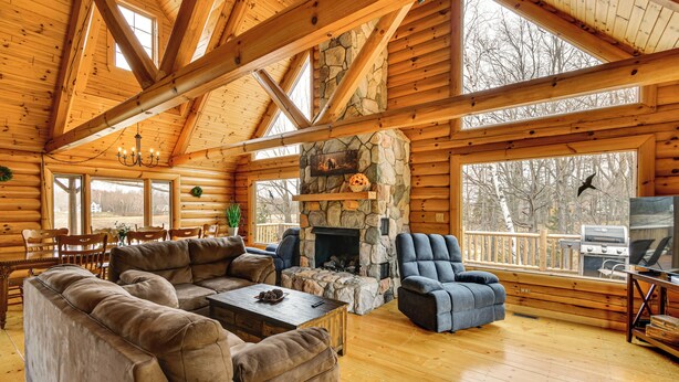 A log cabin interior with a stone fireplace, wooden furniture, and large windows. - Group Fun on Tittabawassee River! Large Log Cabin (Beaverton)