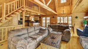 A log cabin interior with a wooden staircase, a large sectional sofa, a dining table, and a kitchen area. - Group Fun on Tittabawassee River! Large Log Cabin (Beaverton)