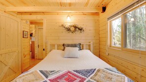A wooden cabin bedroom with a bed, a window, a door, and a wall-mounted light fixture. - Log Cabin Near Dragon Trail at Hardy Dam! (White Cloud)