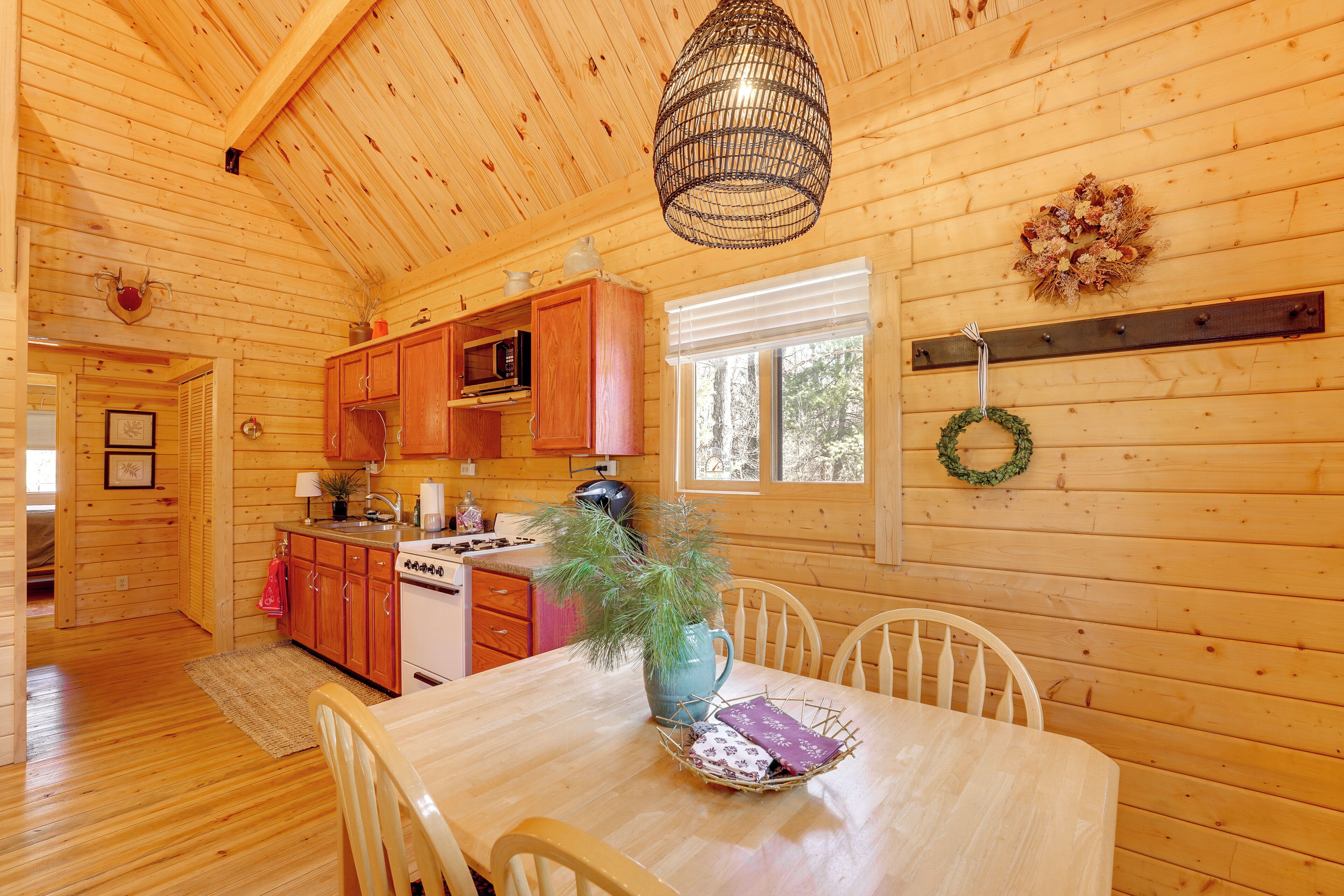 Interior de una cabaña de madera con una mesa de comedor, sillas, zona de cocina y una lámpara colgante.