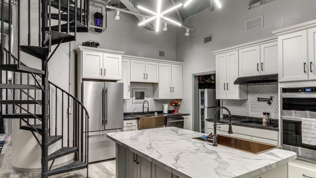 A modern kitchen with white cabinets, a marble island, stainless steel appliances, and a black spiral staircase.
