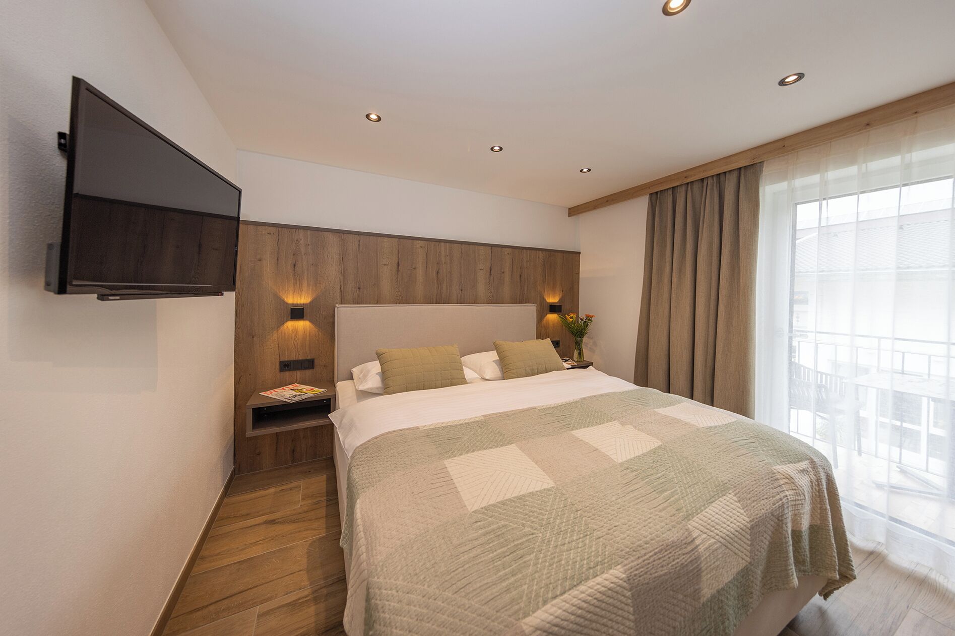 A hotel room with a bed, a TV mounted on the wall, a nightstand, and a window with curtains.