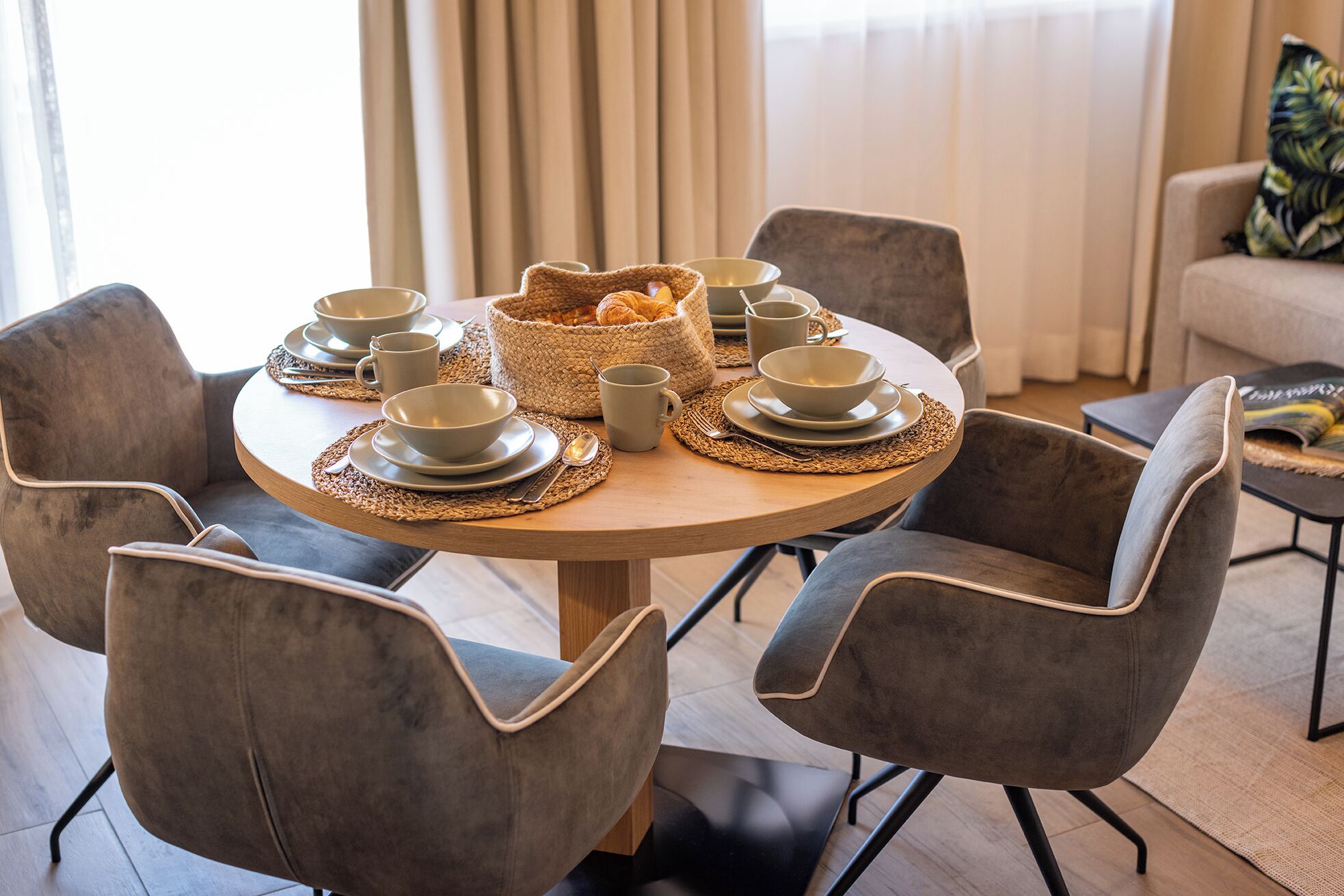 A round dining table with grey upholstered chairs, a woven placemat, and a basket of croissants.
