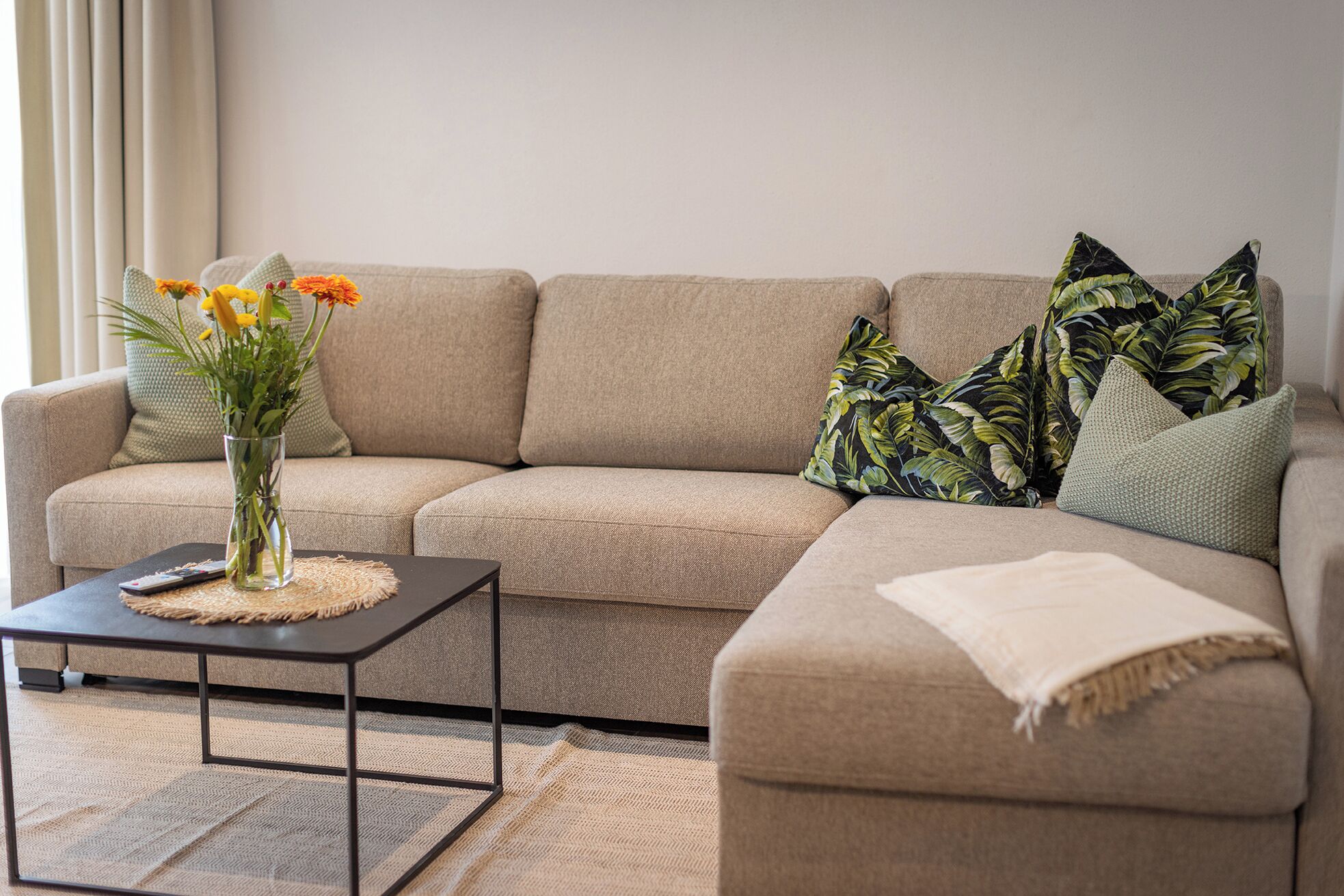 A beige sectional sofa with a vase of yellow flowers on a black coffee table, a remote control, and a woven placemat.