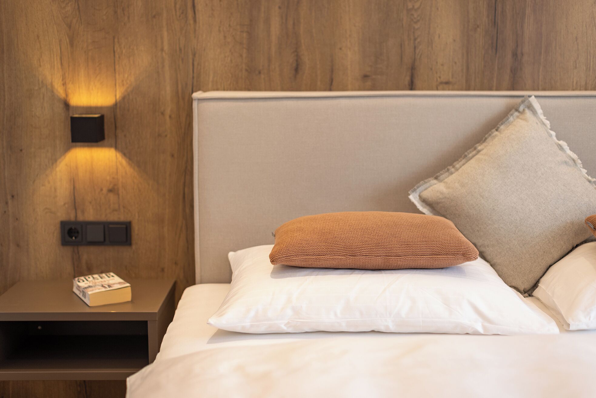 Un lit bien fait, avec du linge de lit blanc, un coussin marron tissé et un coussin beige. On aperçoit une table de chevet avec un livre et une lampe. Le mur derrière le lit est en bois.