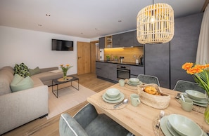 A modern living room with a sofa, coffee table, and dining table set with plates, bowls, and a basket of bread. - Lennis Apartments by we rent (Zell am See)