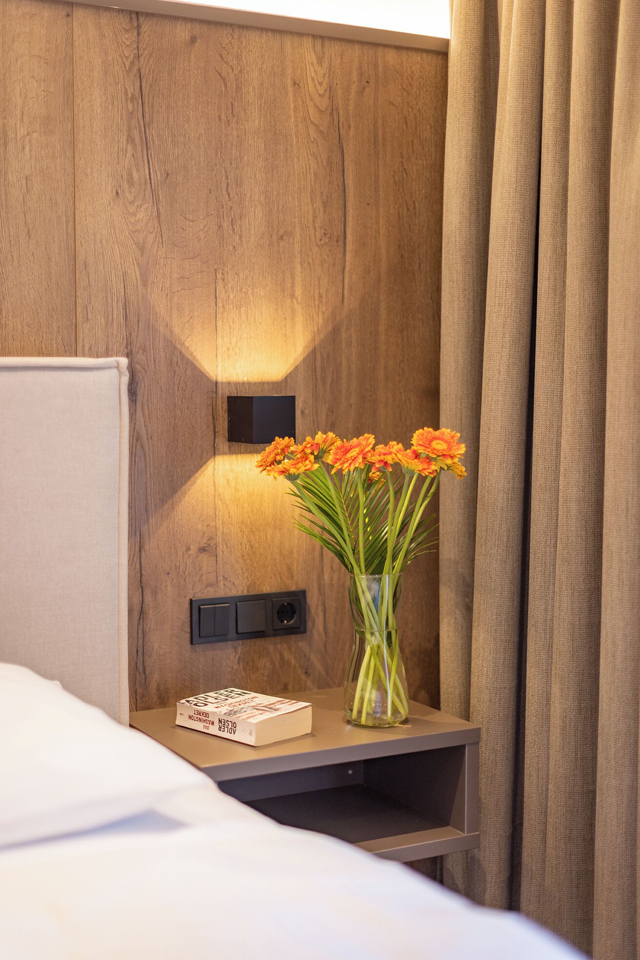 A hotel room with a wooden wall, a bedside table with a book, a vase with flowers, and a curtain.