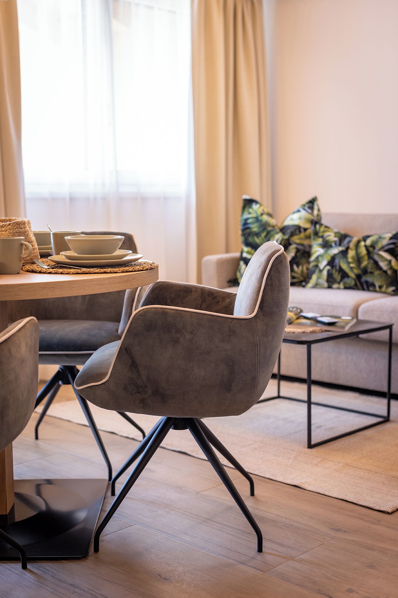 Een moderne woonkamer met een eethoek, met een grijze beklede fauteuil met metalen poten, een houten eettafel en een bank met gestreepte kussens.