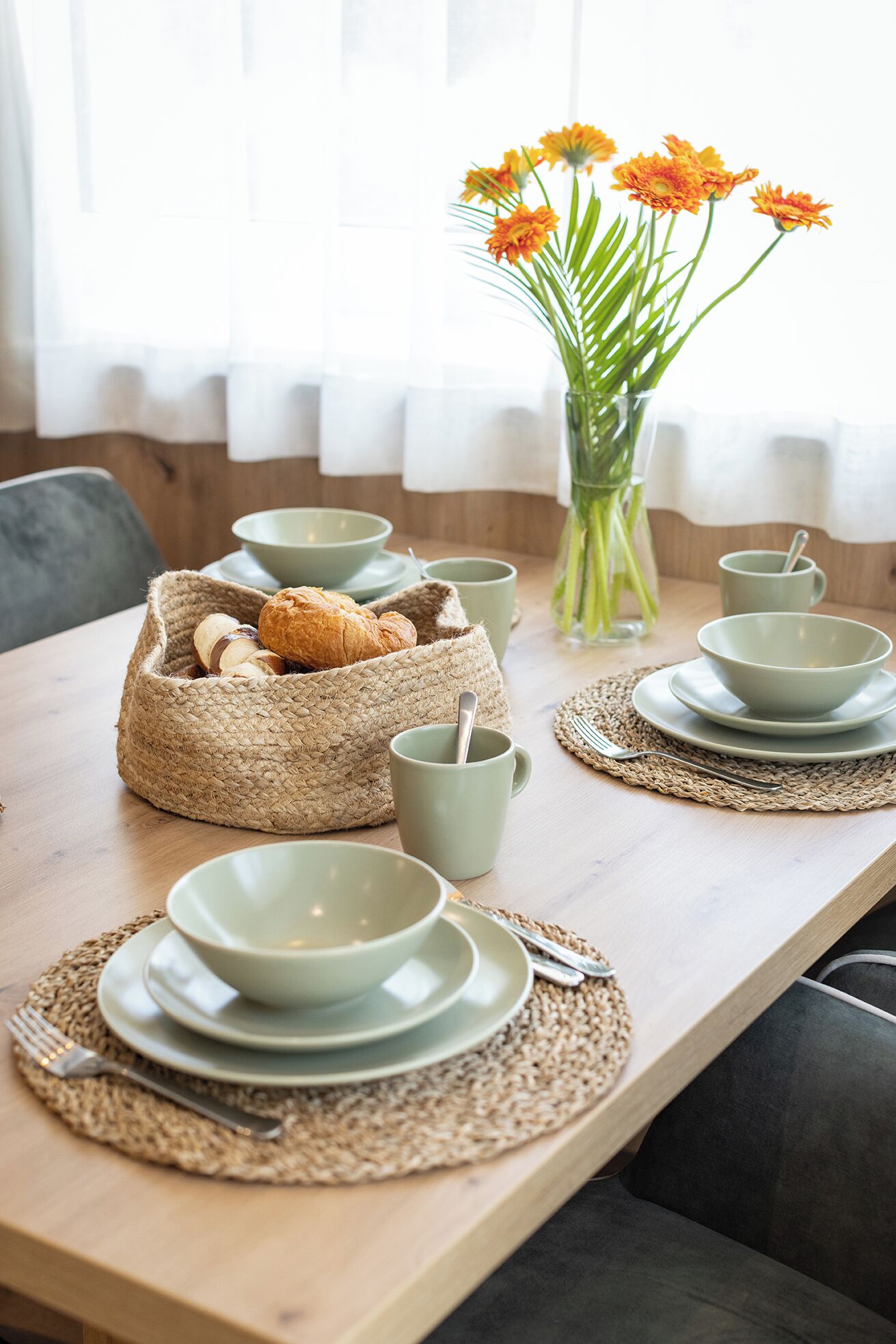 A dining table set with plates, bowls, and a woven basket containing bread.