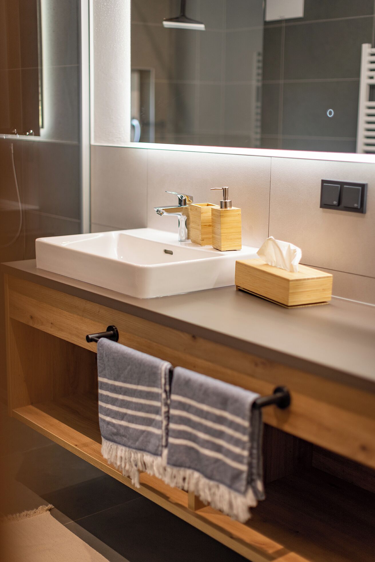 A modern bathroom with a white sink, a wooden vanity, a towel rack, and a mirror.