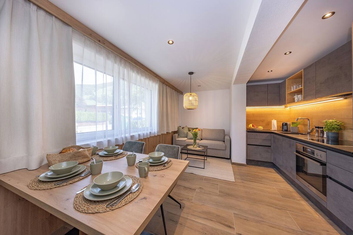 Una cocina moderna con horno empotrado, un comedor con mesa de madera dispuesta para cuatro personas y una zona de estar acogedora con sofá.