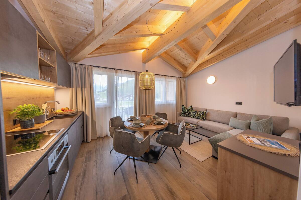 Una cocina moderna con vigas de madera, un comedor con mesa y sillas redondas, un sofá y un televisor montado en la pared.