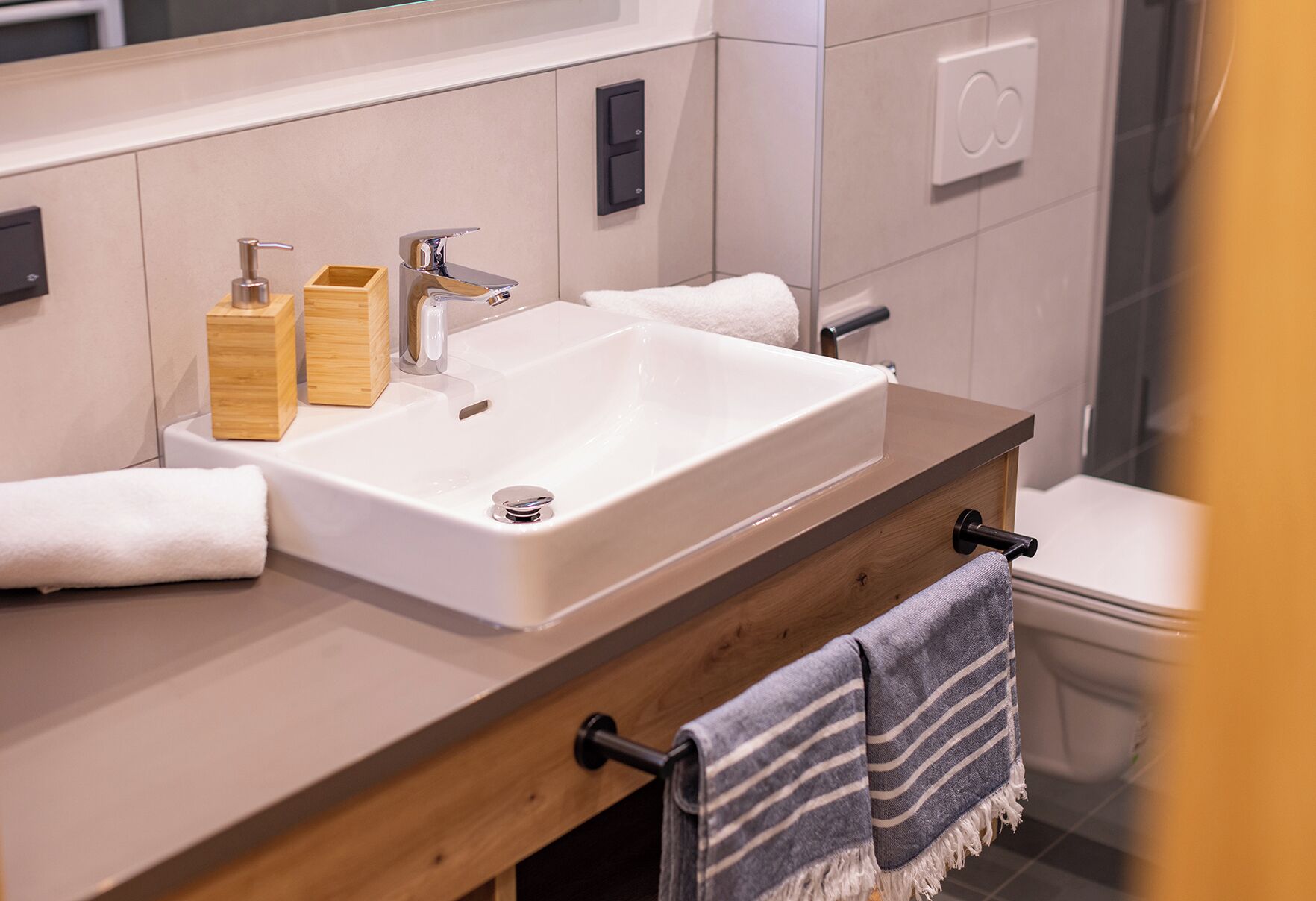 Un baño moderno con un lavamanos grande de color blanco, dispensadores de jabón de madera, un perchero para toallas y un inodoro.