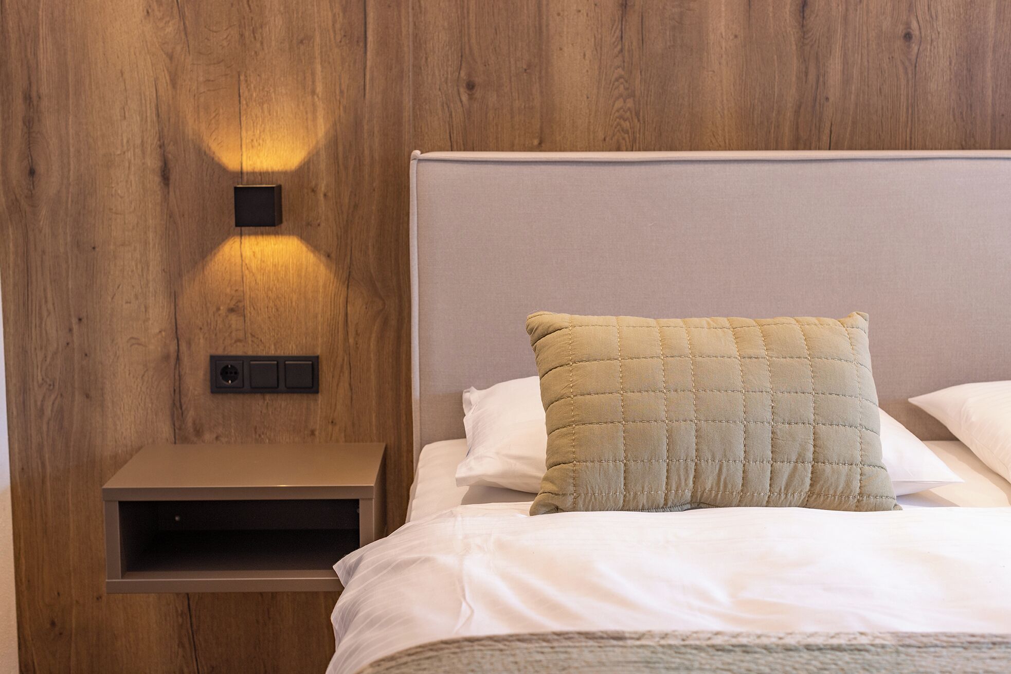 A neatly made bed with a quilted pillow, a bedside table with a lamp, and a wall-mounted switchboard.