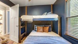 Una cama individual superpuesta con cabecera de madera, una cama inferior con manta azul, y una ventana con persianas.