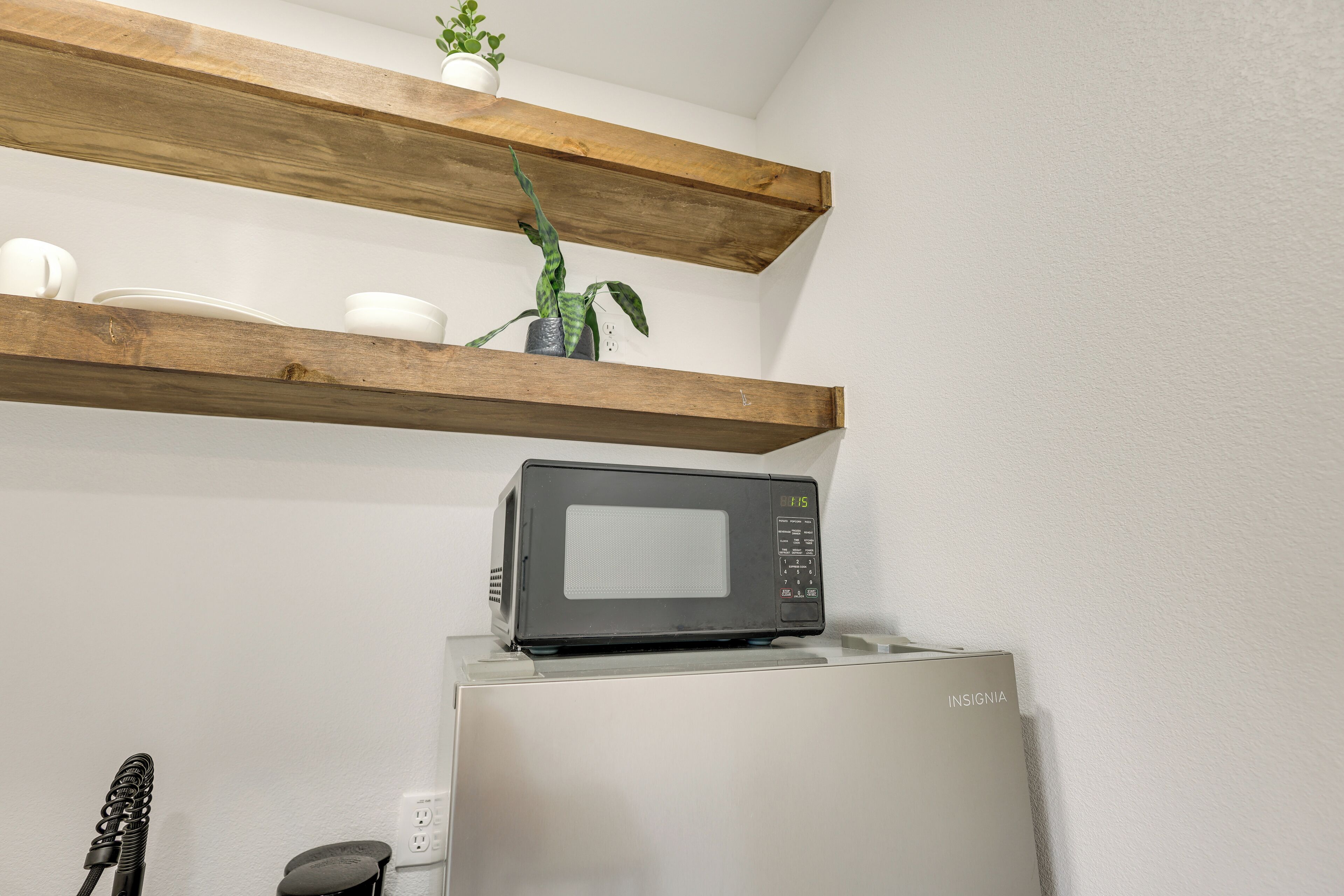 A kitchen with a microwave on top of a refrigerator, wooden shelves above holding dishes and a plant.
