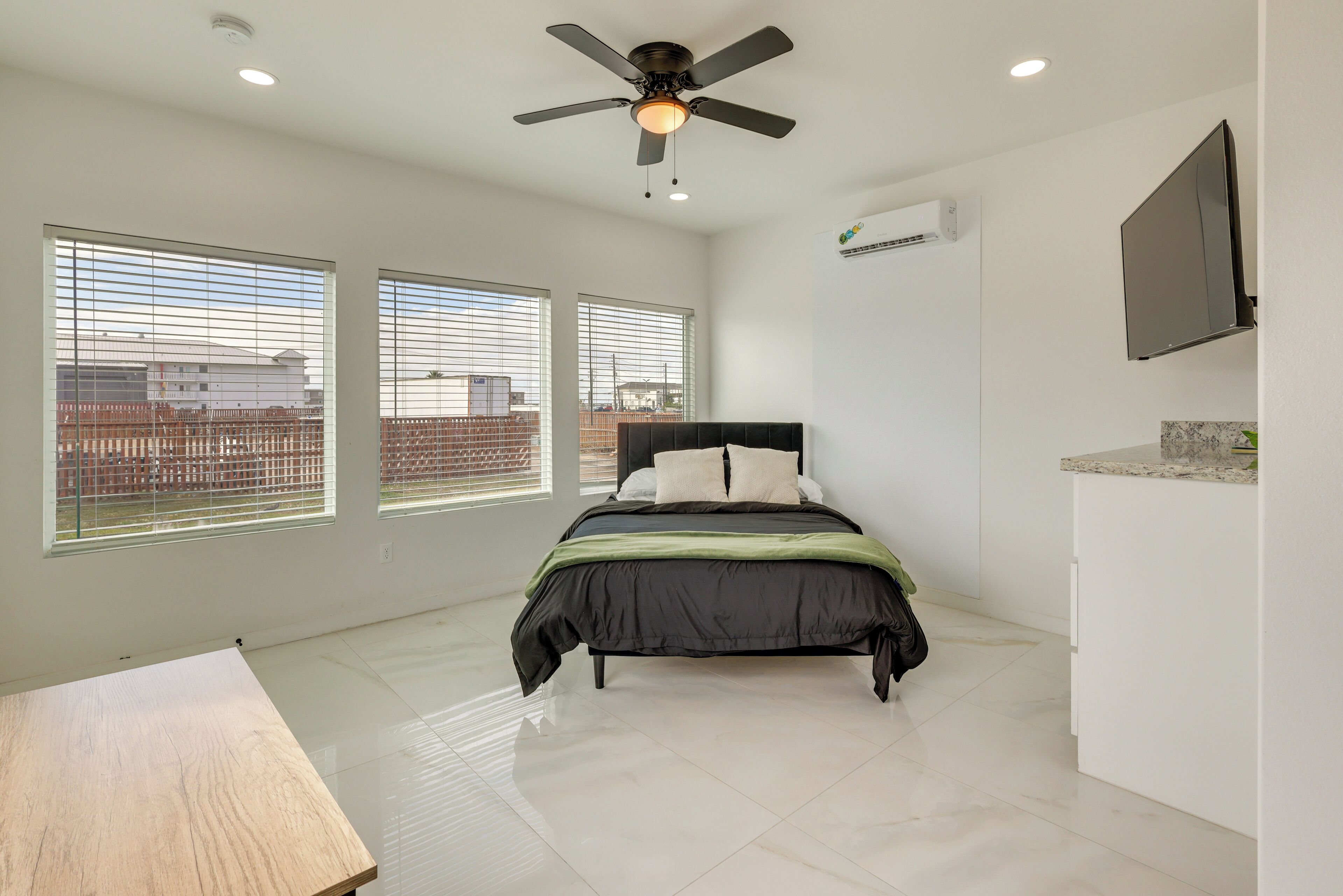 A bedroom with a bed, a ceiling fan, a TV, and large windows.