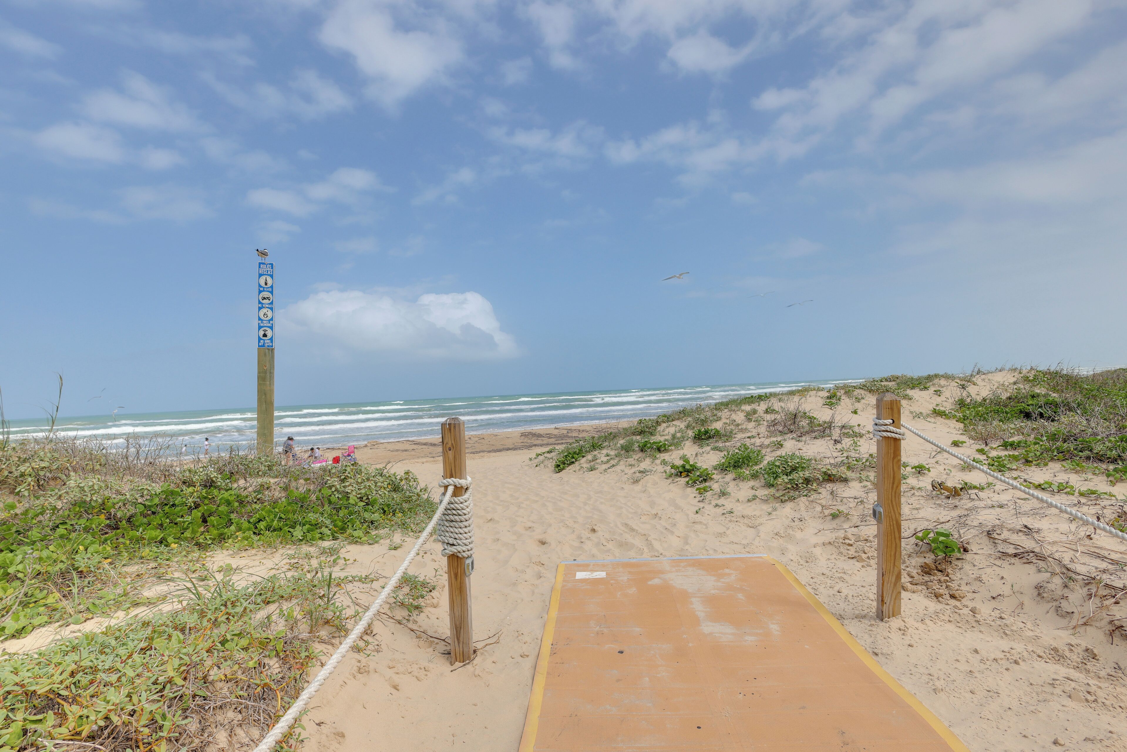 Un sentier de sable qui longe la plage et mène à l’océan, avec un poteau en bois et une barrière faite de cordes.