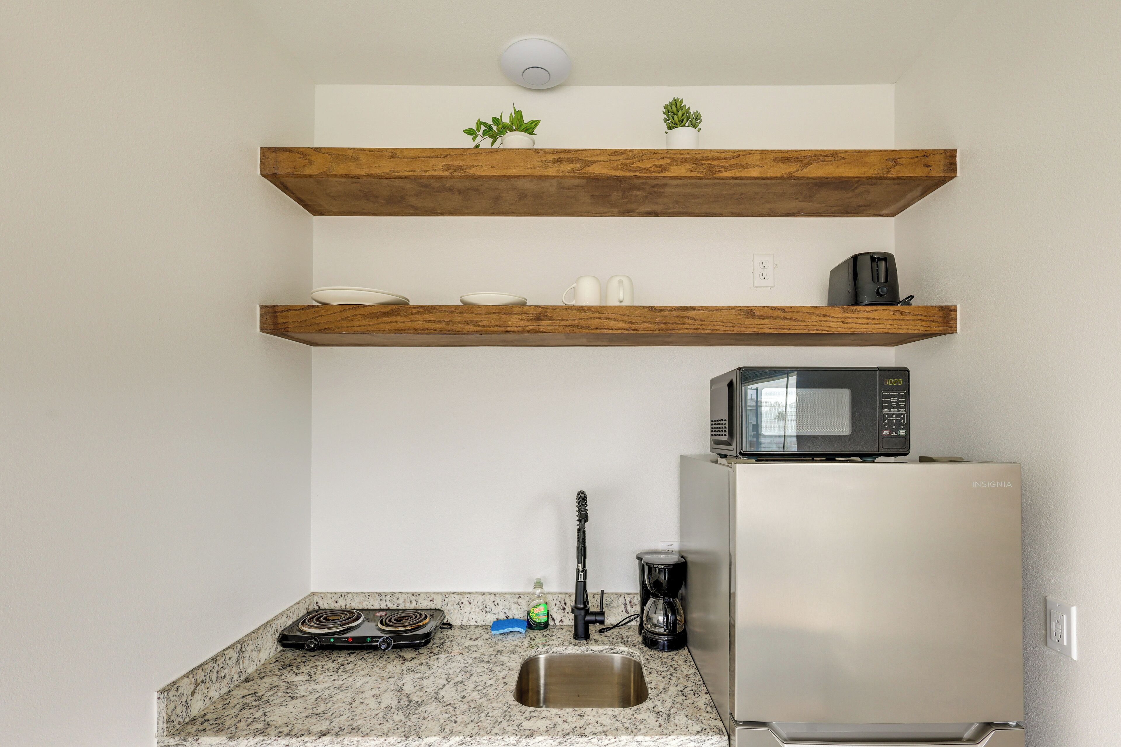 Een keuken met een roestvrijstalen koelkast, een magnetron, een koffiezetapparaat en een spoelbak. Boven het aanrecht zijn houten planken met planten.